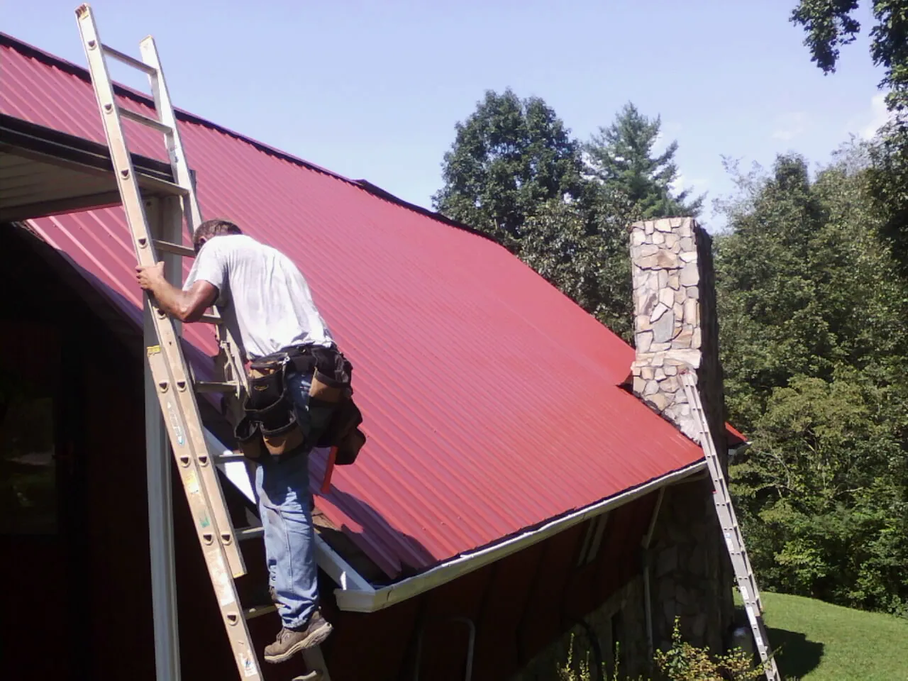 Roofer on a ladder beside a red metal roof and stone chimney — Soffit & Fascia Repair by Roofing Brothers in Orford